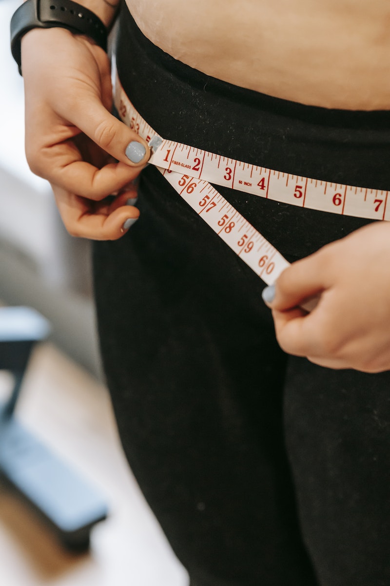 From above of crop anonymous plump female using measuring tape around hips in gym